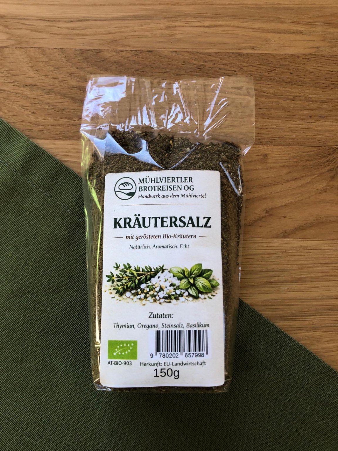 Kräutersalz mit gerösteten Bio-Kräutern in der transparenten 150-g-Packung auf Holz und grünem Stoff